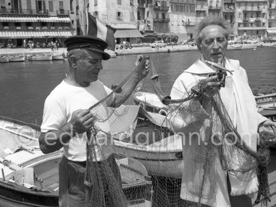On the docks of the harbor at Villefranche-sur-Mer Jean Cocteau receives in a fishnet a life-size gold sardine from fisherman Paul Dunan (left), known as \'The Pirate\'. This present, created by a Nicois jeweller, is from the fishermen as a thank-you for the restoration and decoration works undertaken by the writer on the Chapelle Saint Pierre. Villefranche-Sur-Mer 1959. - Photo by Edward Quinn
