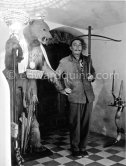 At the entrance of his home, in the "Bear Lobby", Salvador Dalí poses with one of his collection of crossbows and an elaborately decorated bear. Portlligat, Cadaqués, 1957. - Photo by Edward Quinn