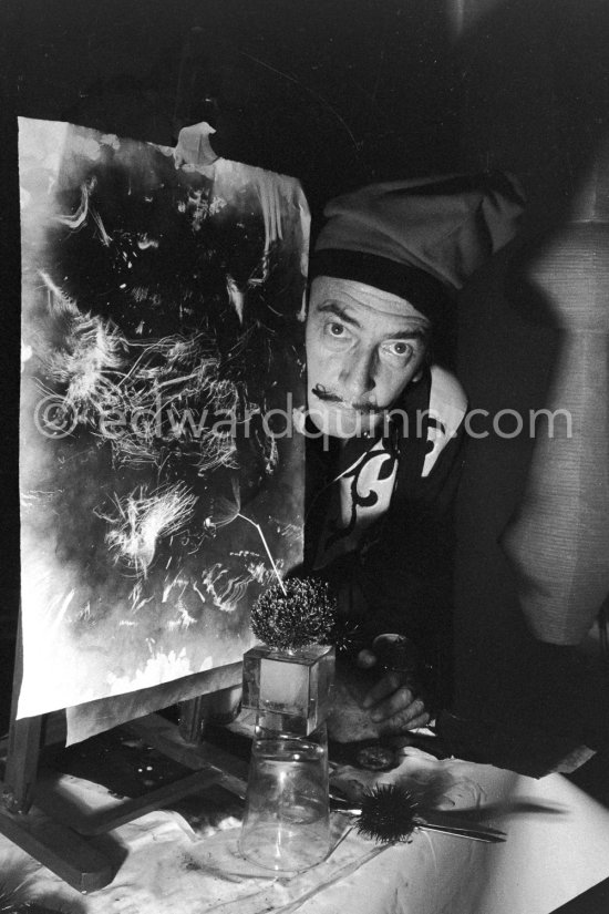 Salvador Dalí shows the set up from which he hopes to obtain the painting, a sea urchin is placed on an overturned inkwell glass and in its mouth, the"Aristotle's lantern", which has the movements of a human hand) he puts a swan’s feather or here a light dried up flower. The object is allowed to slightly touch a sheet of blackened paper. The movements then made by the sea urchin are traced on the paper. At Salvador Dalí's house, Portlligat, Cadaqués 1957. - Photo by Edward Quinn