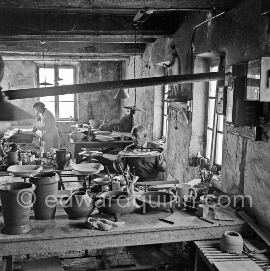 Pablo Picasso with the plate "Le picador". Suzanne Ramié (in the background) at work at the Madoura pottery in Vallauris. Above him is a statue of St-Claude, the patron saint of the potters, whose name was given to Pablo Picasso\'s younger son. Vallauris 23.3.1953. - Photo by Edward Quinn