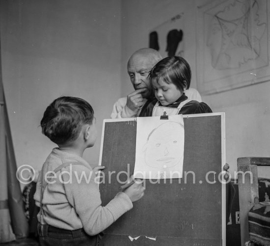 Encouraged by his father, Claude Picasso tries to do a portrait of Pablo Picasso, portraying him with a round face, deep furrows on his forehead, a triangle for a nose, and streaks of hair on the sides of his head. La Galloise, Vallauris 16.4.1953. - Photo by Edward Quinn