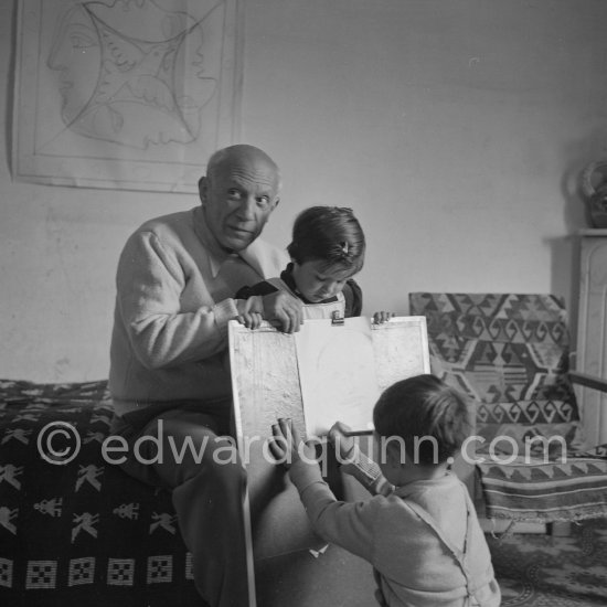 Encouraged by his father, Claude Picasso tries to do a portrait of Pablo Picasso, portraying him with a round face, deep furrows on his forehead, a triangle for a nose, and streaks of hair on the sides of his head. La Galloise, Vallauris 16.4.1953. - Photo by Edward Quinn
