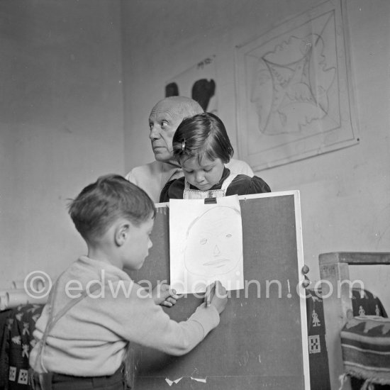 Encouraged by his father, Claude Picasso tries to do a portrait of Pablo Picasso, portraying him with a round face, deep furrows on his forehead, a triangle for a nose, and streaks of hair on the sides of his head. La Galloise, Vallauris 16.4.1953. - Photo by Edward Quinn