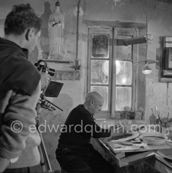 Pablo Picasso using a sharp instrument to scrape the surface of a clay plate as he designs a portrait. During filming of "Pablo Picasso", director Luciano Emmer, (left) and Pablo Picasso. With a statue of St-Claude Picasso, the patron saint of the potters, whose name was given to his younger son. Madoura pottery, Vallauris 14.10.1953. - Photo by Edward Quinn