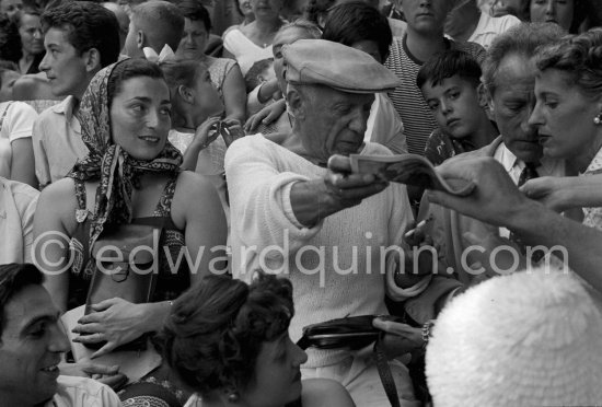 On the grandstand of a bullfight put on in his honor, Pablo Picasso is signing autographs. On the left of Pablo Picasso Jacqueline, on the right Jean Cocteau and Francine Weisweiller, behind Pablo Picasso partly hidden his children Paloma Picasso and Claude Picasso and Gérard Sassier. Vallauris 1955. - Photo by Edward Quinn