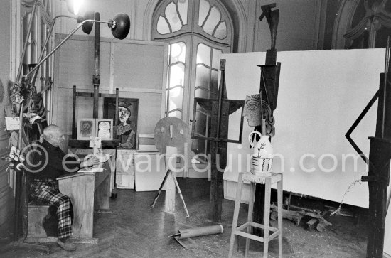 Pablo Picasso sitting at a school bench. Dressed in the checkered trousers that amuse him because of their association with the self-portraits of Courbet, Pablo Picasso toys with a small cardboard sculpture. La Californie, Cannes 1957. - Photo by Edward Quinn