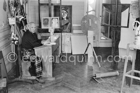 Pablo Picasso sitting at a school bench. Dressed in the checkered trousers that amuse him because of their association with the self-portraits of Courbet, Pablo Picasso toys with a small cardboard sculpture. La Californie, Cannes 1957. - Photo by Edward Quinn