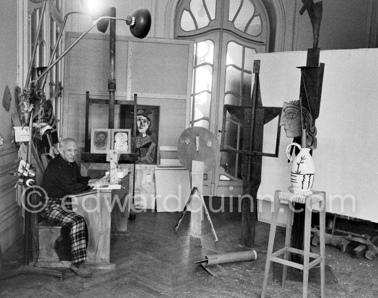 Pablo Picasso sitting at a school bench. Dressed in the checkered trousers that amuse him because of their association with the self-portraits of Courbet, Pablo Picasso toys with a small cardboard sculpture. La Californie, Cannes 1957. - Photo by Edward Quinn