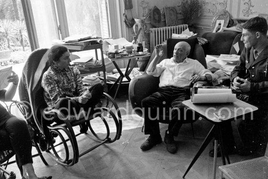 Pablo Picasso and Jacqueline and an unidentified soldier. Jacqueline, sitting in a rocking chair, is wearing a blouse made from a textile designed by Pablo Picasso. A table by Joseph-Marius Tiola in the background. La Californie, Cannes 1961. - Photo by Edward Quinn