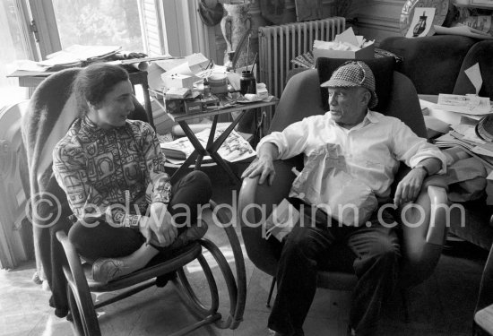 Pablo Picasso and Jacqueline. He never fails to surprise with the unusual, in this case a check dear-stalker worn with all the aplomb of a 19th century English Squire. Jacqueline, sitting in a rocking chair, is wearing a blouse made from a textile designed by Pablo Picasso. A table by Joseph-Marius Tiola in the background. La Californie, Cannes 1961. - Photo by Edward Quinn