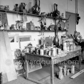 A table with tubs of paint. At bottom left, some paintings inspired by Paloma Picasso and Claude Picasso and on the shelves, some dolls that Pablo Picasso painted on bits of wood for Paloma Picasso. Studio Le Fournas, Vallauris 1953. - Photo by Edward Quinn