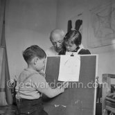 Encouraged by his father, Claude Picasso tries to do a portrait of Pablo Picasso, portraying him with a round face, deep furrows on his forehead, a triangle for a nose, and streaks of hair on the sides of his head. La Galloise, Vallauris 16.4.1953. - Photo by Edward Quinn