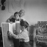 Encouraged by his father, Claude Picasso tries to do a portrait of Pablo Picasso, portraying him with a round face, deep furrows on his forehead, a triangle for a nose, and streaks of hair on the sides of his head. La Galloise, Vallauris 16.4.1953. - Photo by Edward Quinn