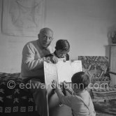Encouraged by his father, Claude Picasso tries to do a portrait of Pablo Picasso, portraying him with a round face, deep furrows on his forehead, a triangle for a nose, and streaks of hair on the sides of his head. La Galloise, Vallauris 16.4.1953. - Photo by Edward Quinn