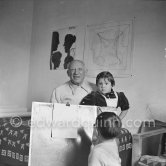 Encouraged by his father, Claude Picasso tries to do a portrait of Pablo Picasso, portraying him with a round face, deep furrows on his forehead, a triangle for a nose, and streaks of hair on the sides of his head. La Galloise, Vallauris 16.4.1953. - Photo by Edward Quinn